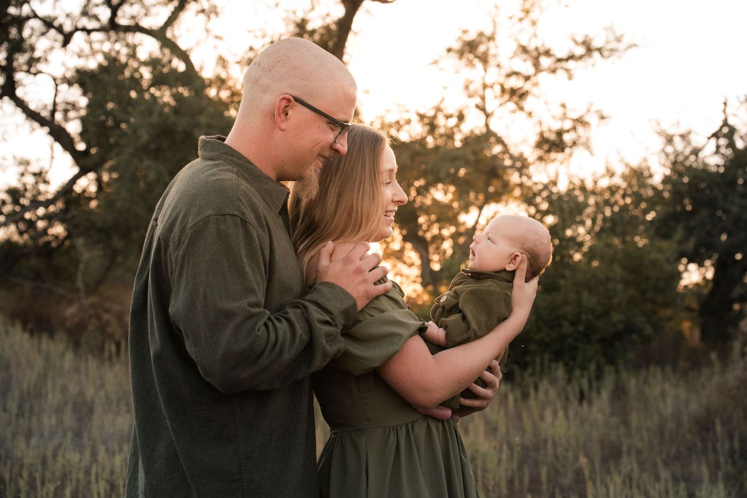 A couple in matching green outfits holds their newborn baby in a golden sunset field.