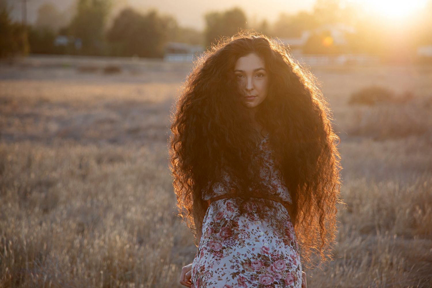 Young woman with long voluminous curly hair wearing a floral dress standing in a golden sunlit field.