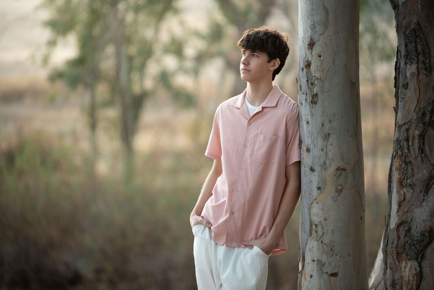 Teenage boy in pink shirt and white pants leaning against a tree trunk in a dry outdoor landscape.