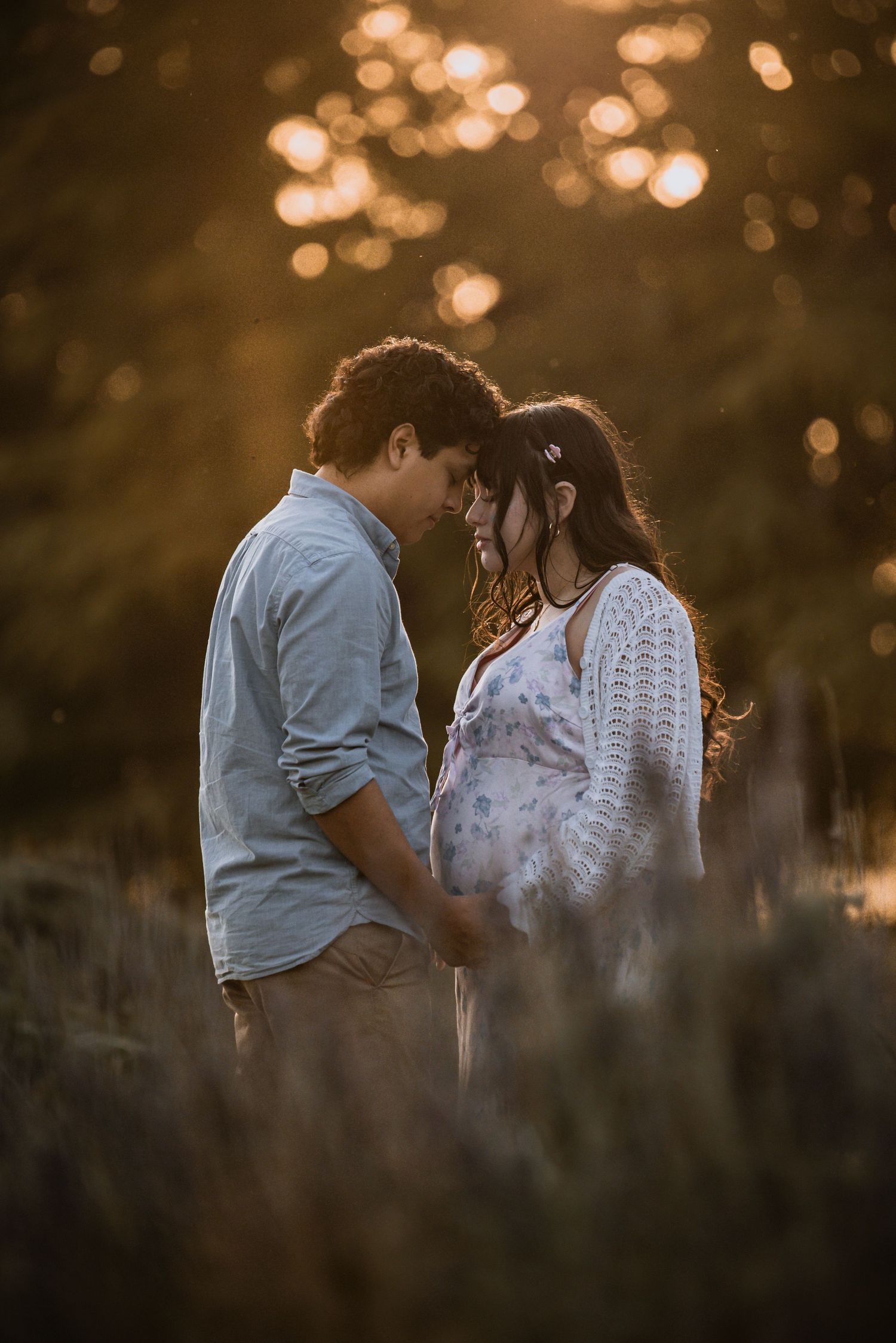 Couple holding hands and touching foreheads tenderly in a golden sunlit lavender field during a maternity photo session.