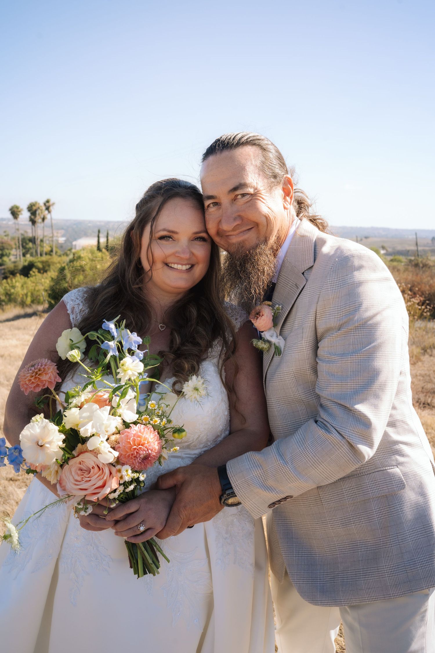 Smiling newlyweds pose together holding bridal bouquet of pink roses at sunny outdoor wedding venue.