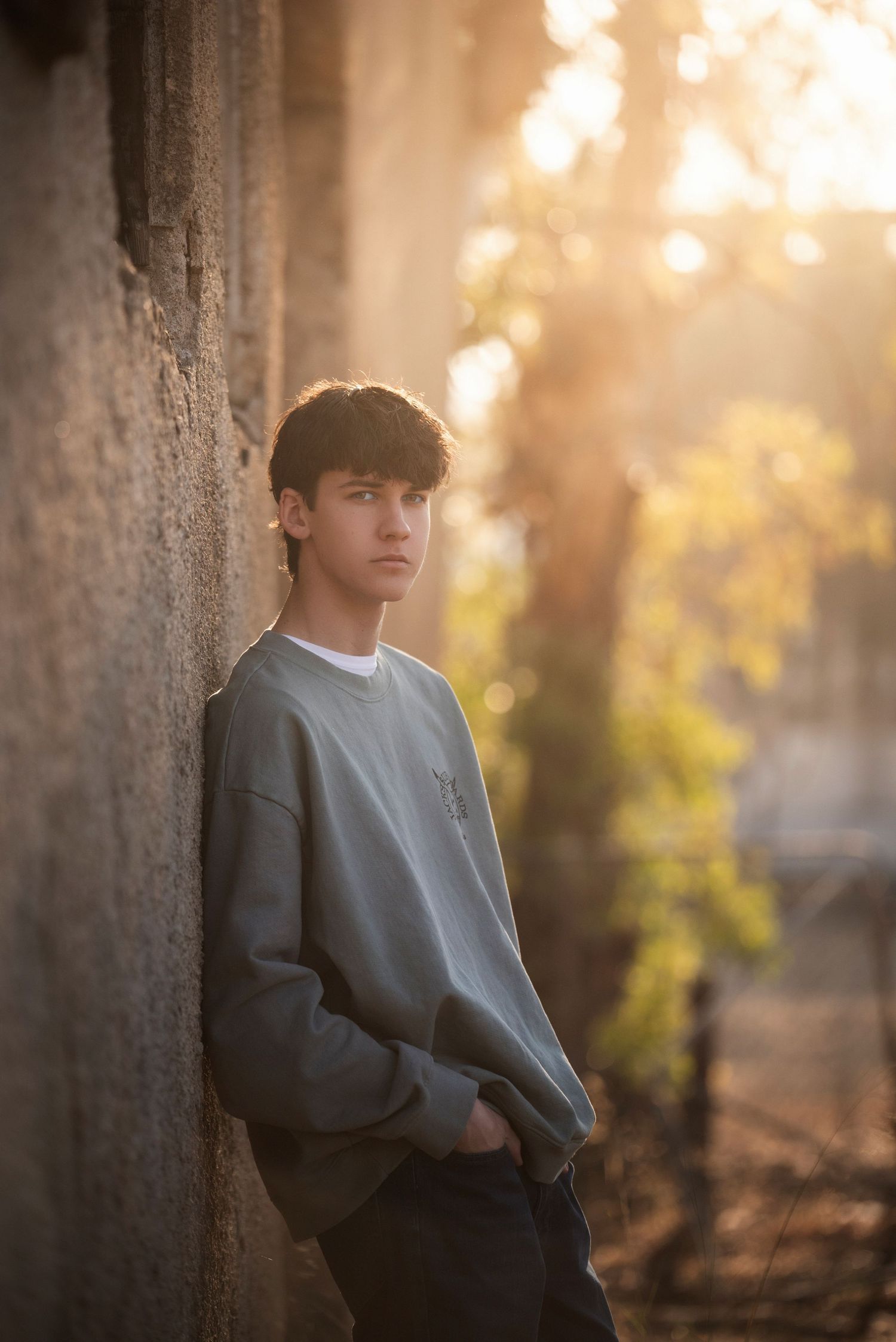 Teenage boy in grey sweatshirt leaning against stone wall in warm golden bokeh sunlight outdoors.