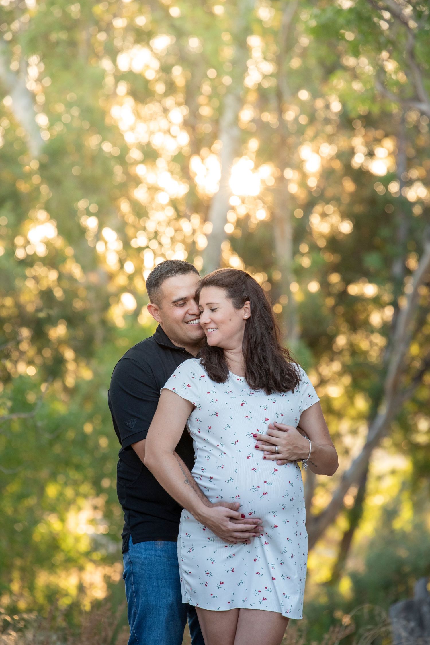 Couple sharing a tender maternity moment outdoors, man kissing woman's cheek while cradling her pregnant belly in golden sunlight.