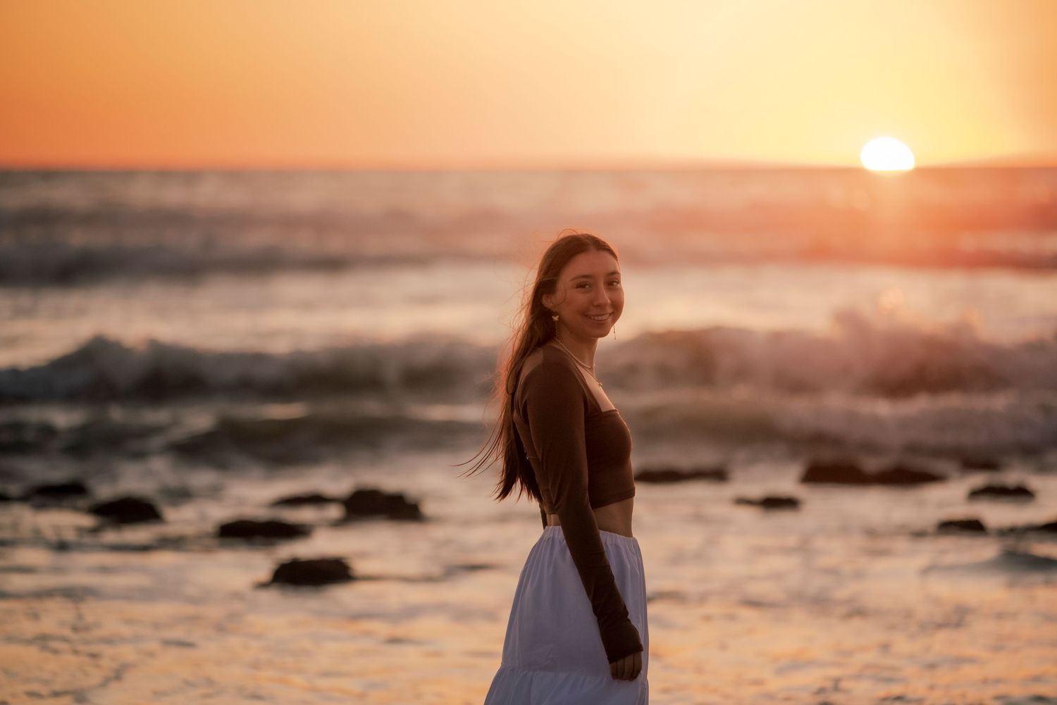 Smiling woman in black top and white skirt standing on beach at golden sunset.