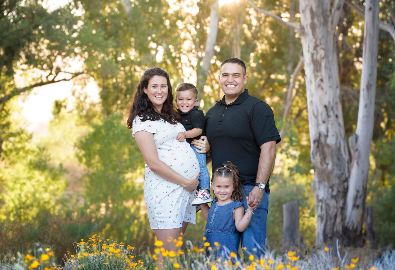 A smiling pregnant family of four poses among wildflowers and eucalyptus trees in warm sunset light.