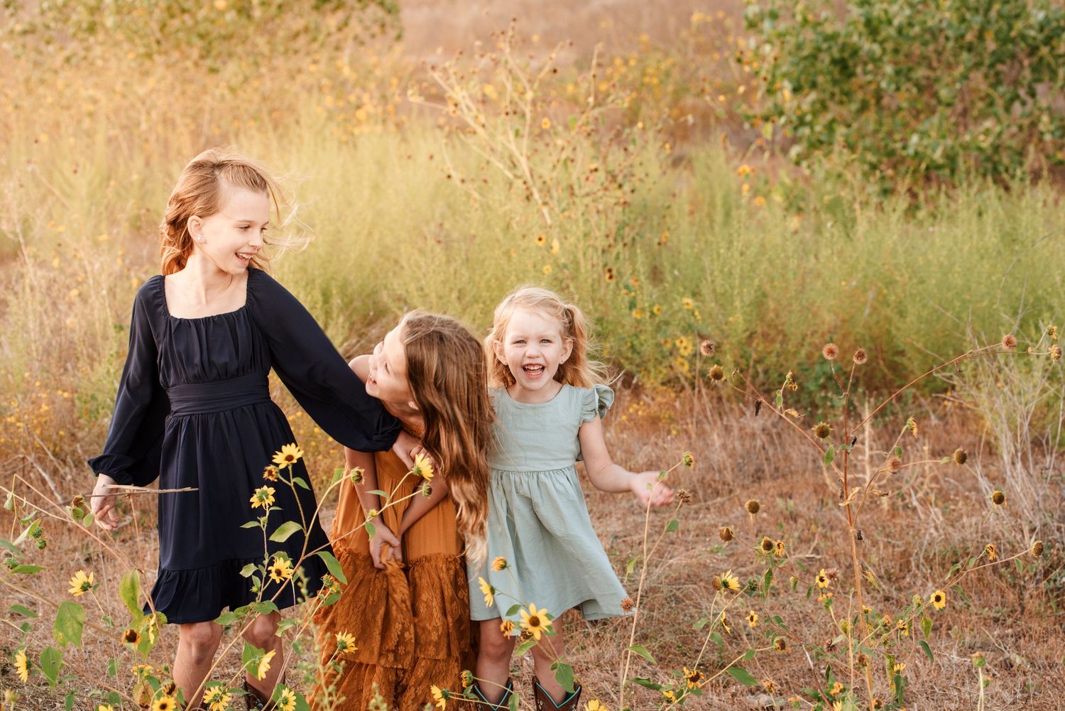 Three little girls in dresses playing and laughing in a sunny meadow.