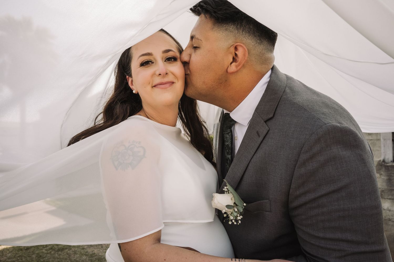 Groom in gray suit kisses brides cheek under flowing white veil in romantic outdoor wedding portrait.