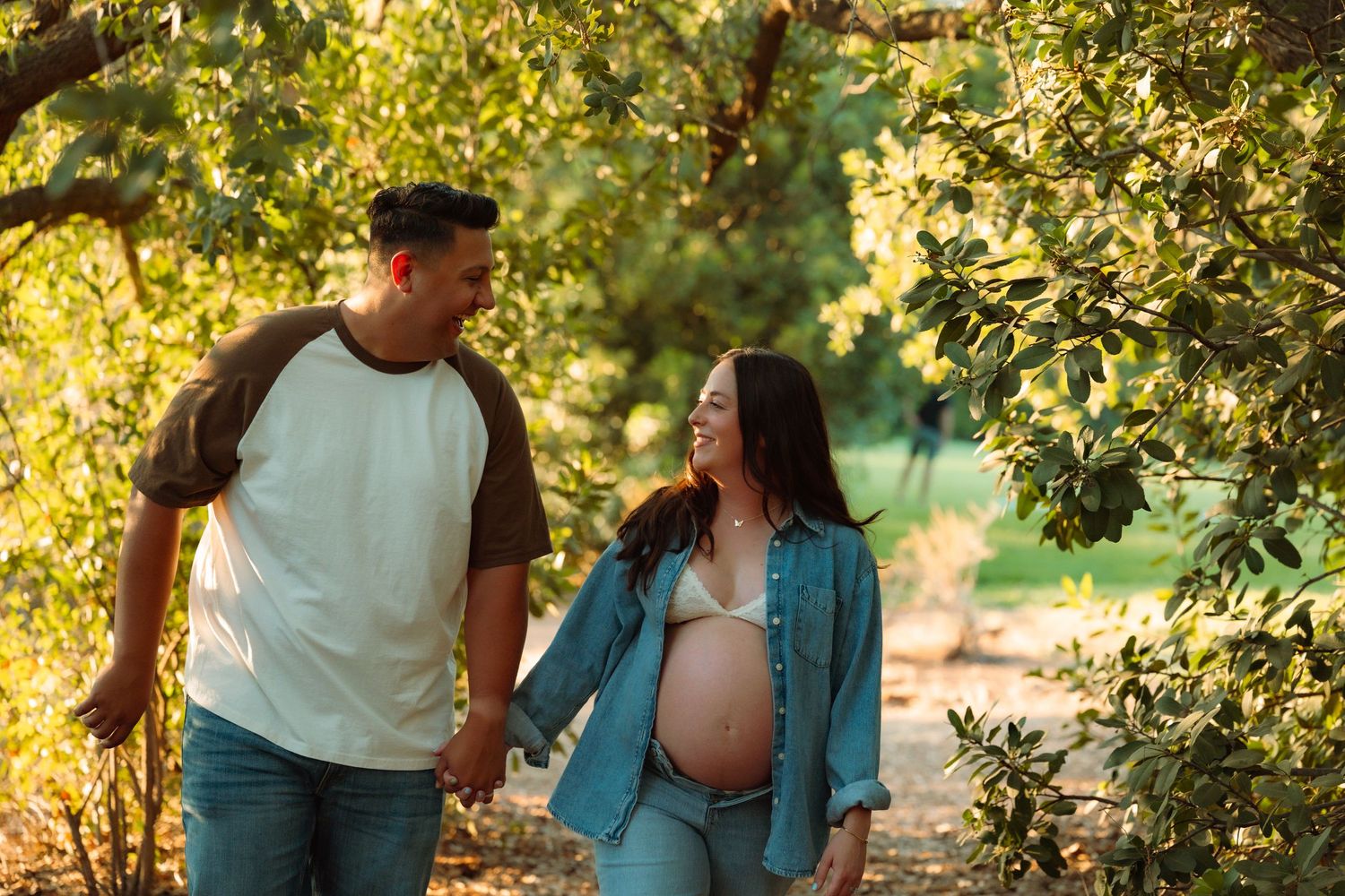 Expectant couple walks hand in hand through sun-dappled trees, smiling at each other during maternity photo session.