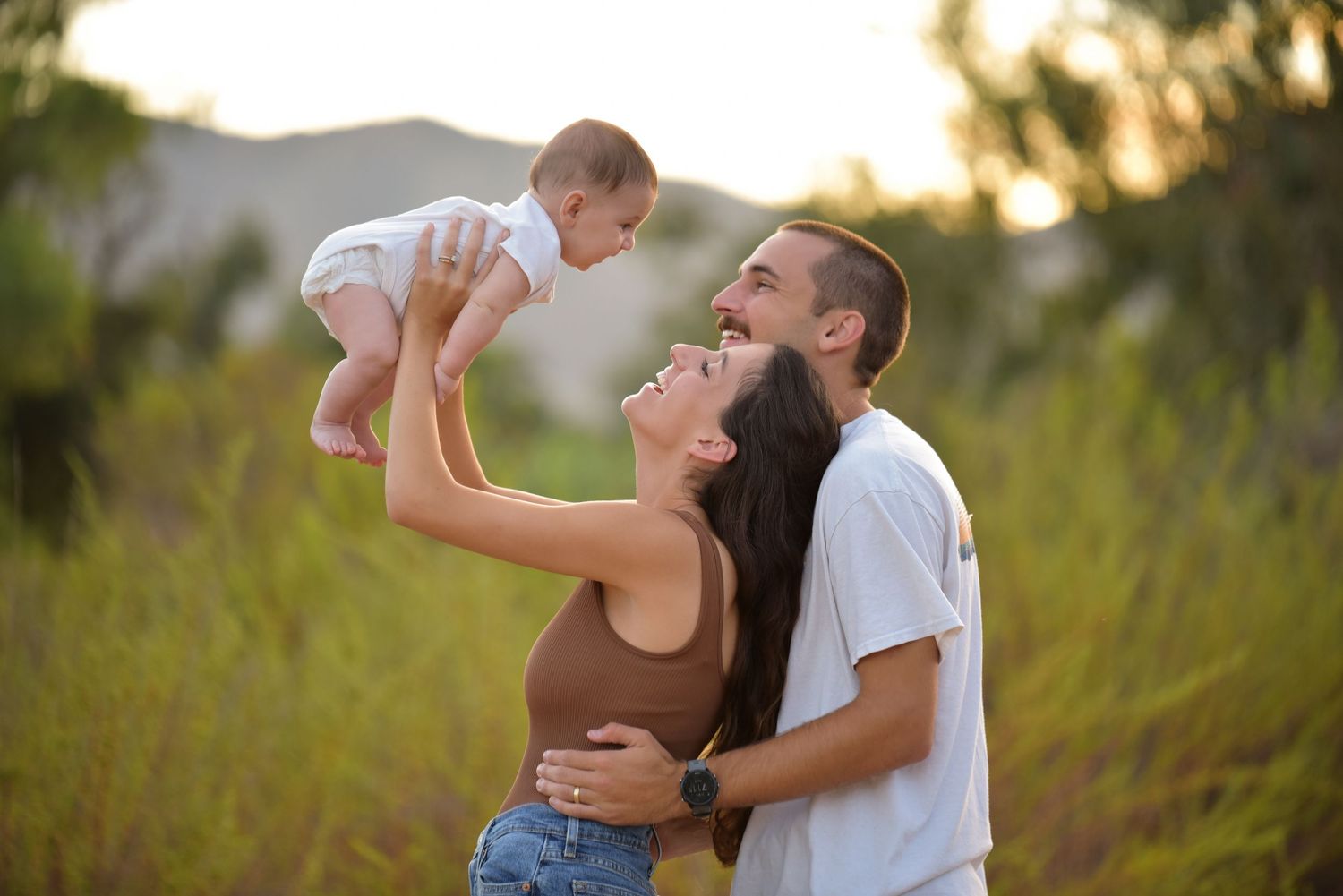 Young parents lifting and laughing with baby in green field at sunset.