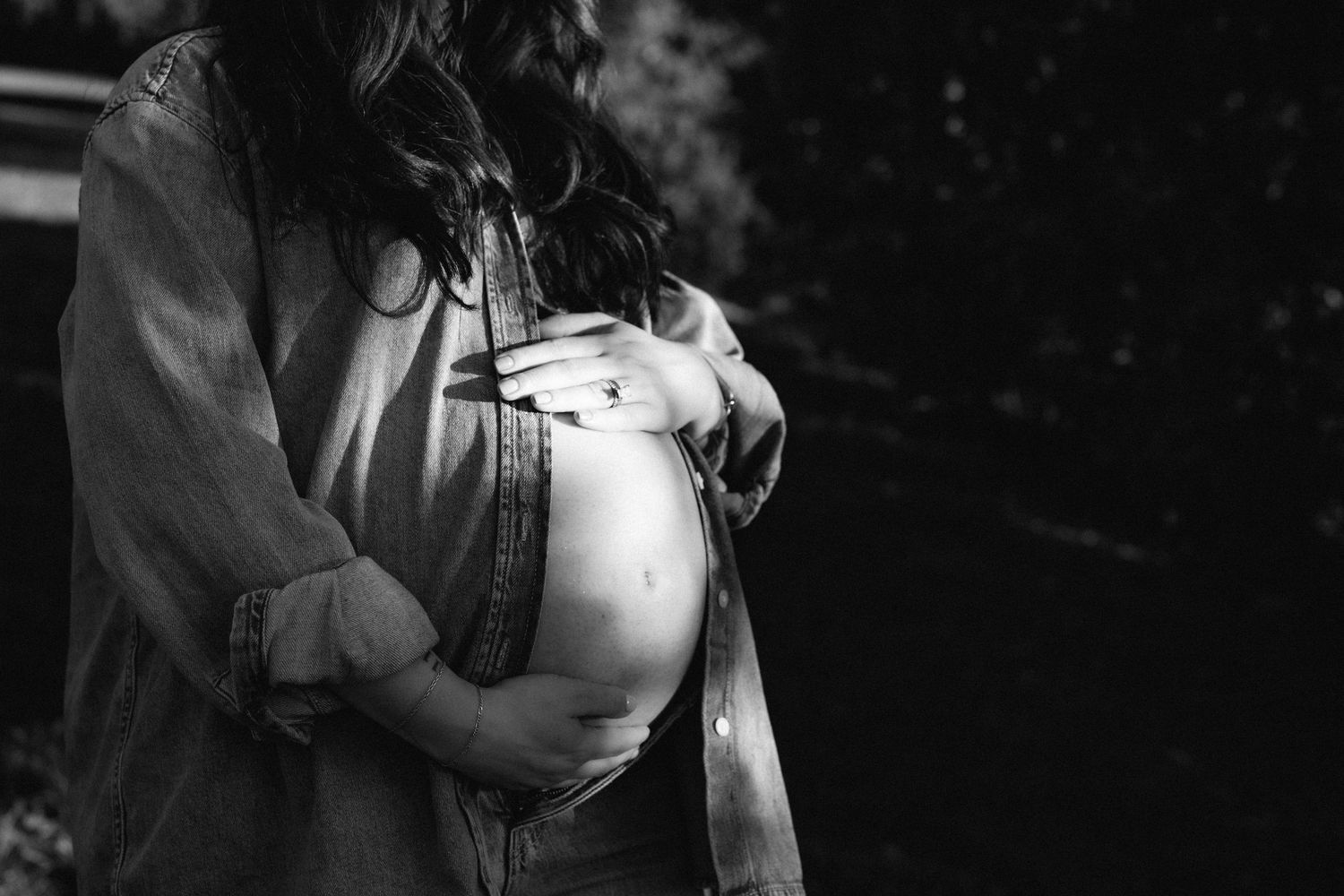 Black and white close-up of pregnant woman cradling her bare baby bump outdoors in soft natural light.