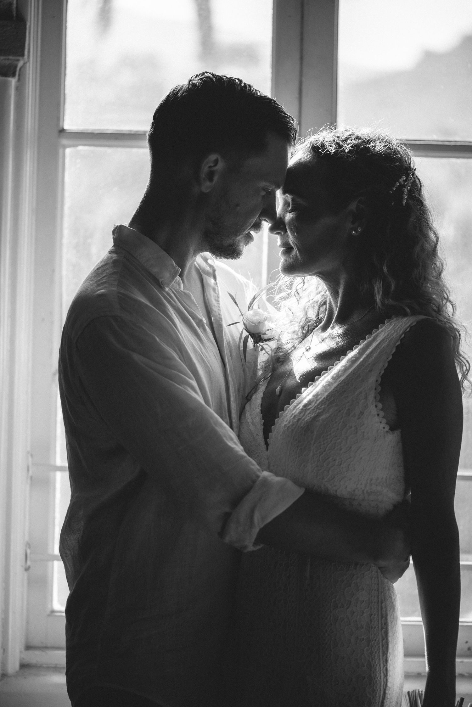 Black and white photo of a romantic couple embracing and facing each other near a sunlit window.