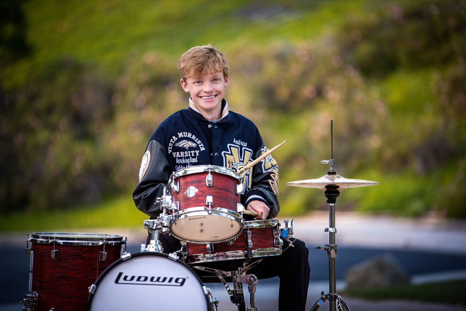 Smiling young boy playing a Ludwig drum kit outdoors on a paved surface with green hills behind.