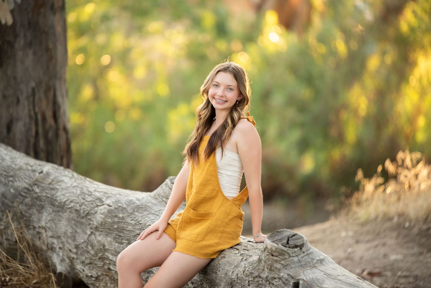 Smiling teenage girl in mustard yellow romper sitting on a fallen log in an autumn forest setting.