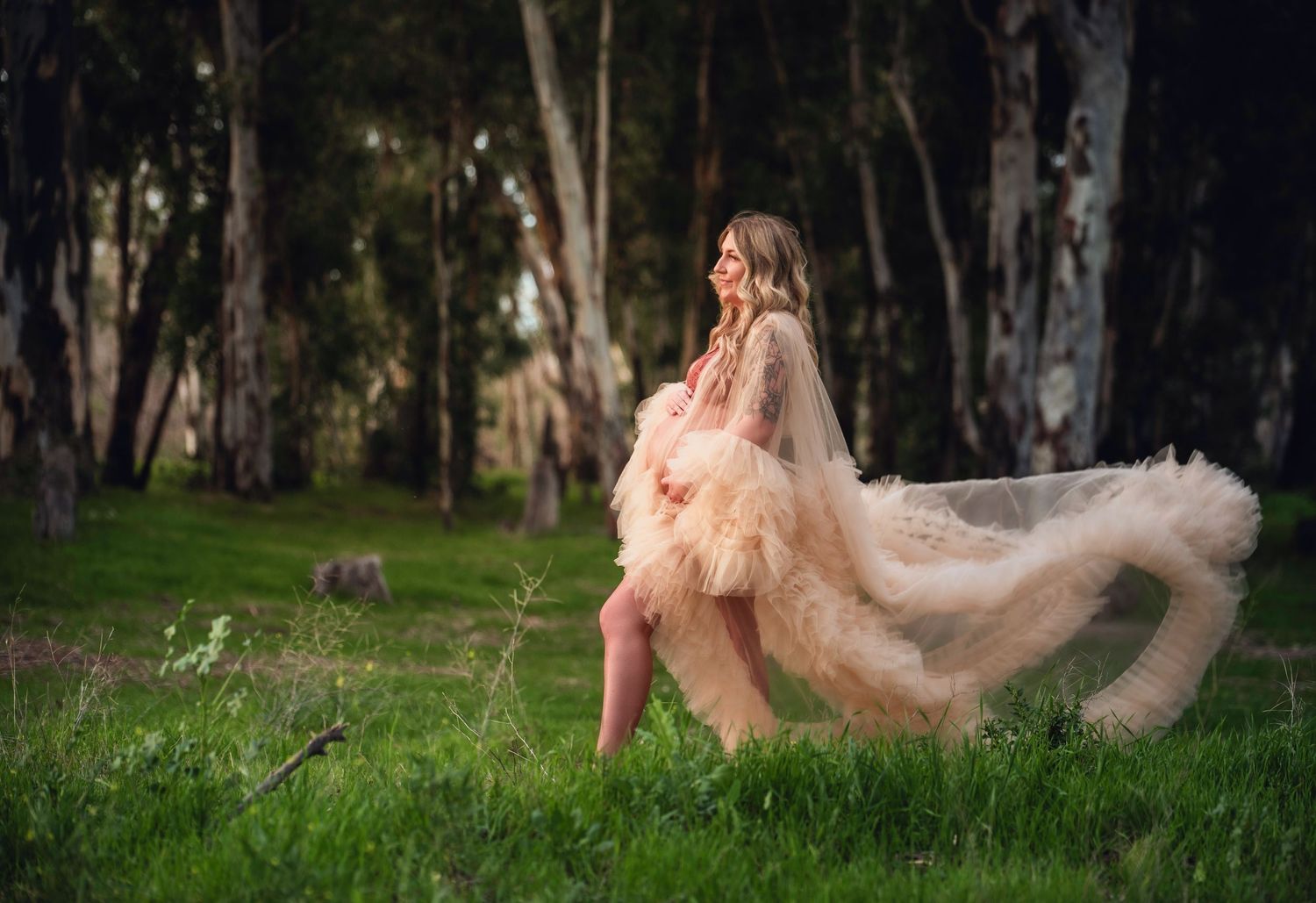 Pregnant woman in flowing champagne tulle robe twirling gracefully in a lush green forest setting.