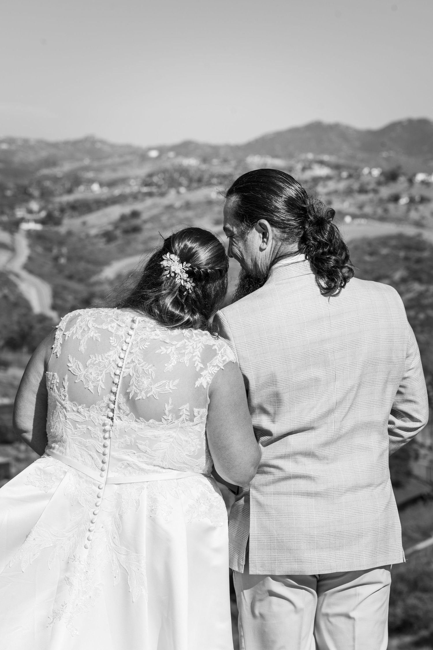 Newlyweds stand back to back overlooking scenic mountain valley landscape in black and white wedding photo.