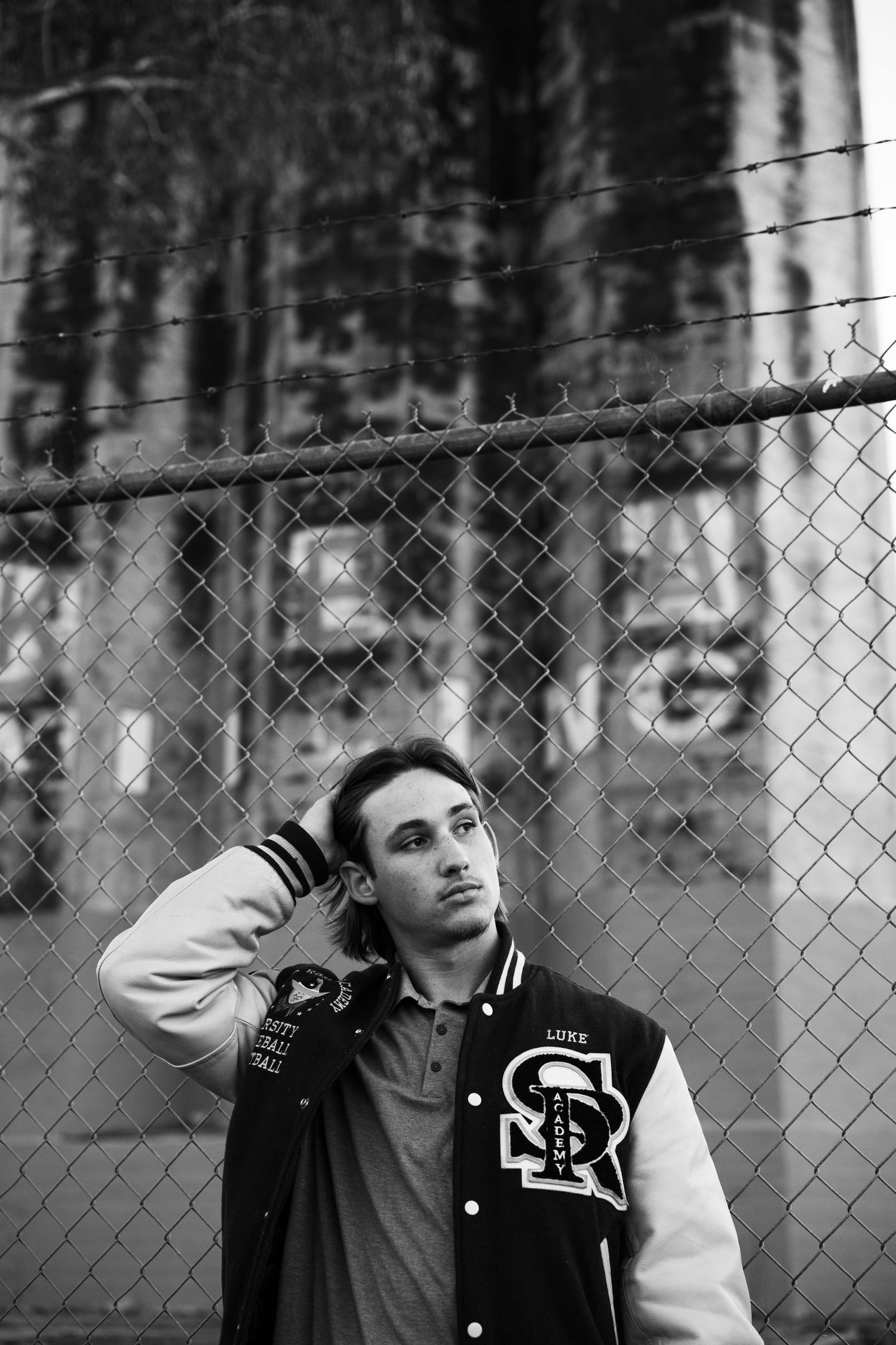 Black and white portrait of young man in varsity jacket leaning against chain-link fence.