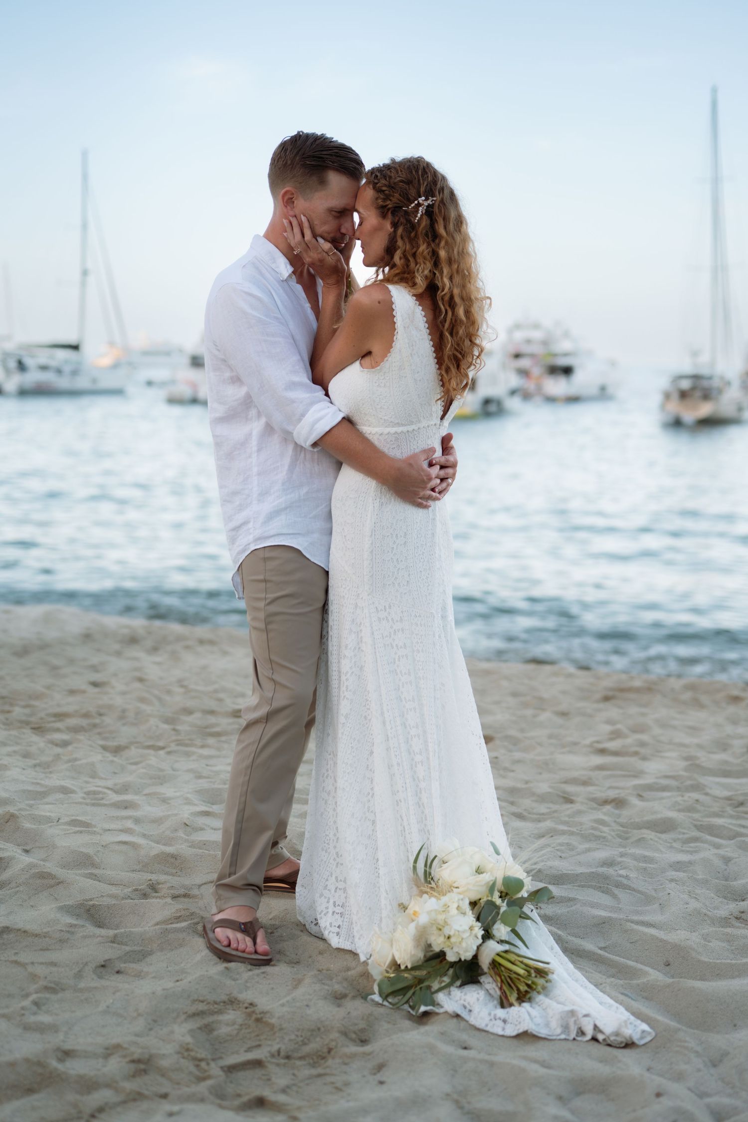 Bride and groom share a romantic kiss on a sandy beach at sunset, with sailboats in the background.
