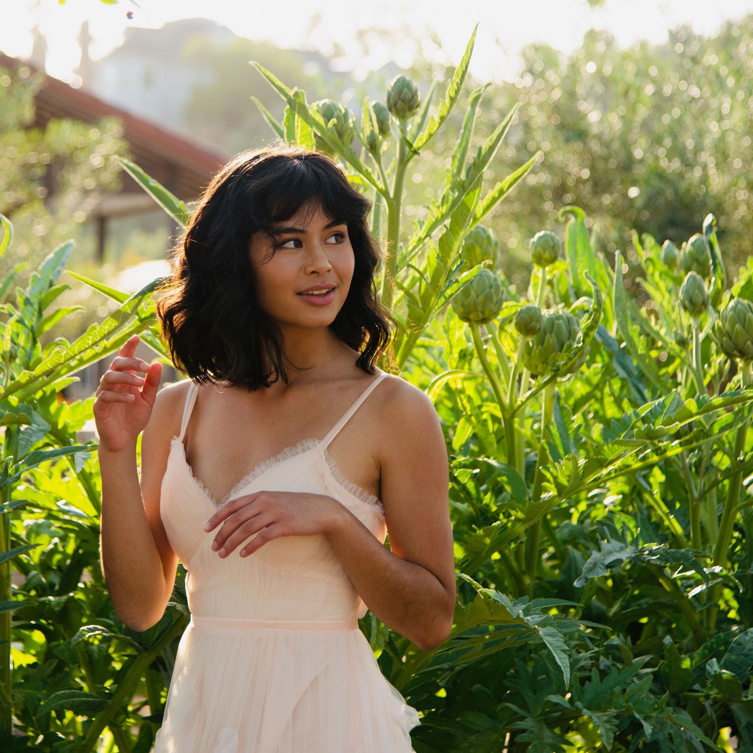 Young woman in blush spaghetti strap dress standing among tall green plants in a sunlit garden.