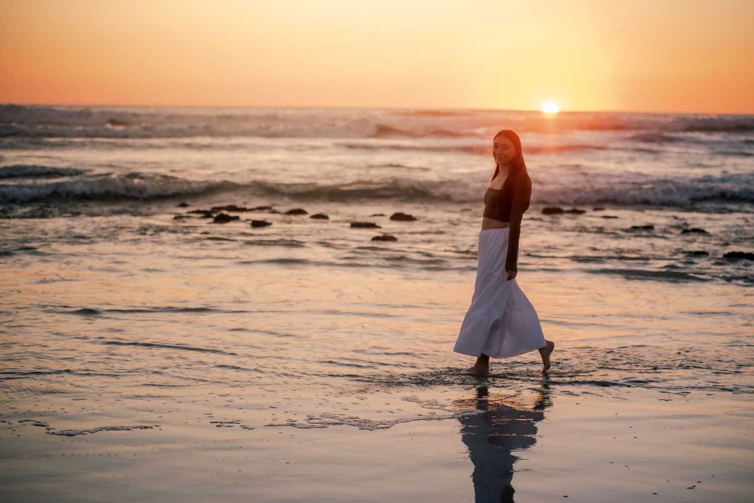 Woman in black top and white skirt walking along ocean shore at vibrant sunset.