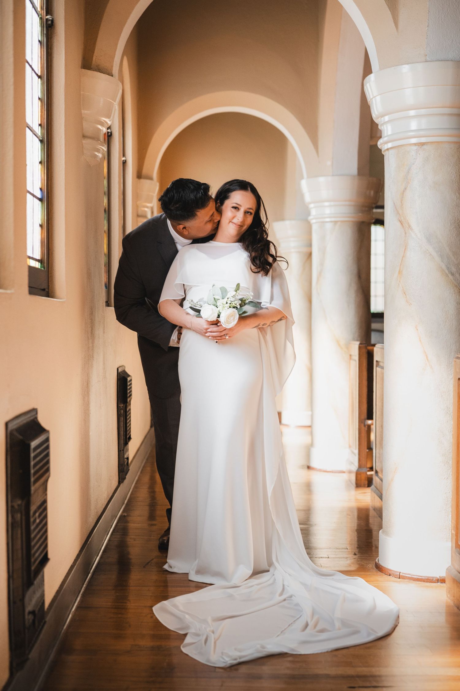 Bride in elegant white cape gown holds bouquet as groom kisses her cheek in a sunlit marble hallway.