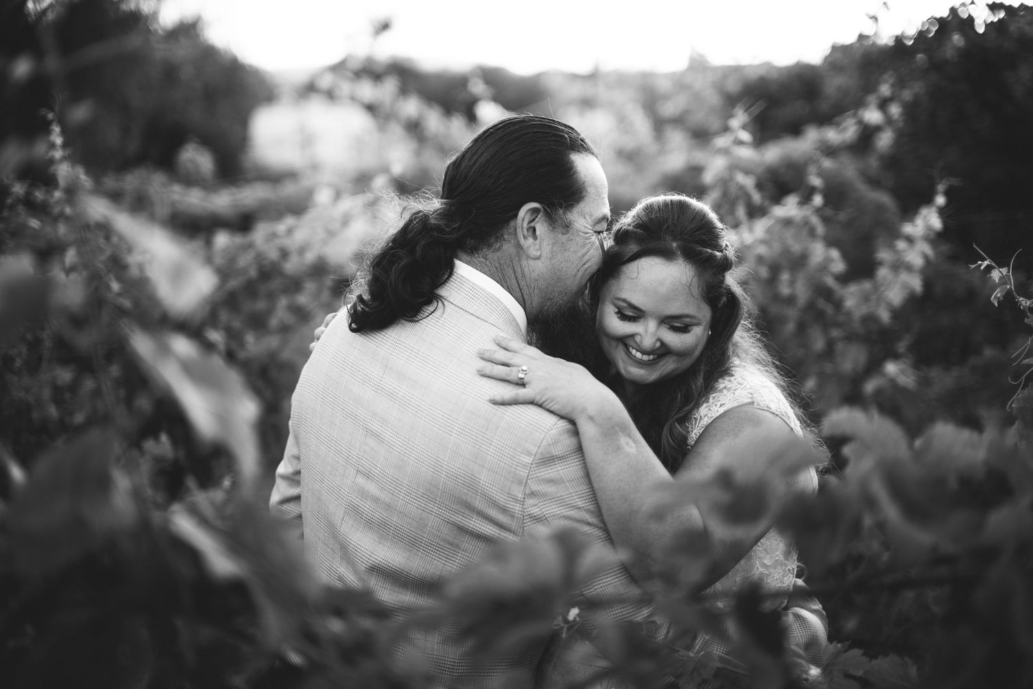 Black and white photo of a smiling couple embracing among leafy vineyard vines, the woman laughing over the man's shoulder.