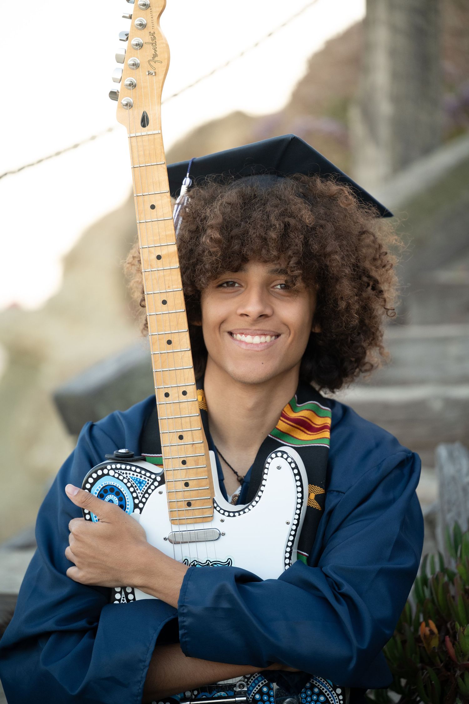 Smiling graduate with curly hair holds electric guitar while wearing cap and gown with colorful kente stole.