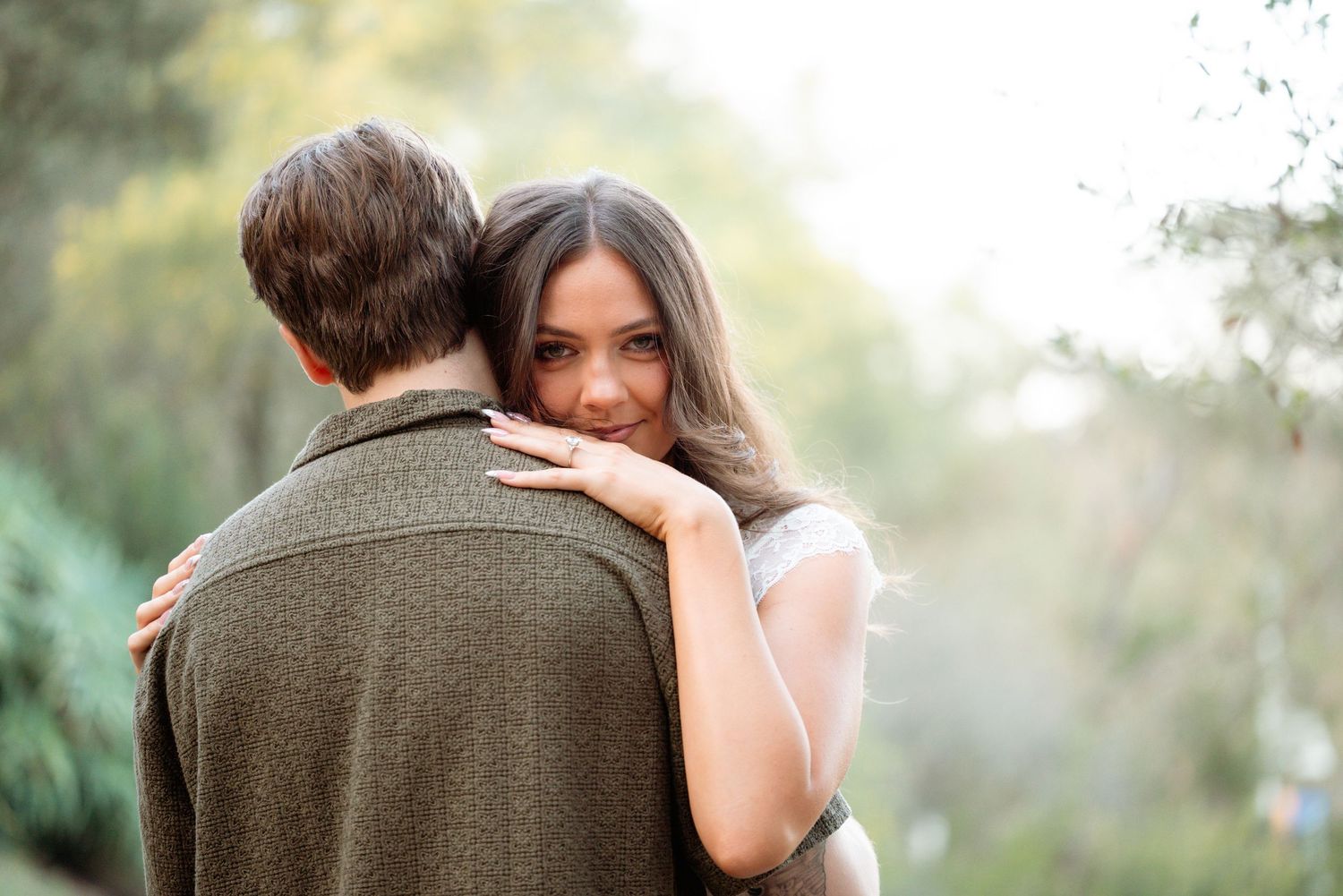 A woman smiles warmly while embracing a man outdoors, resting her head on his shoulder in a lush green setting.