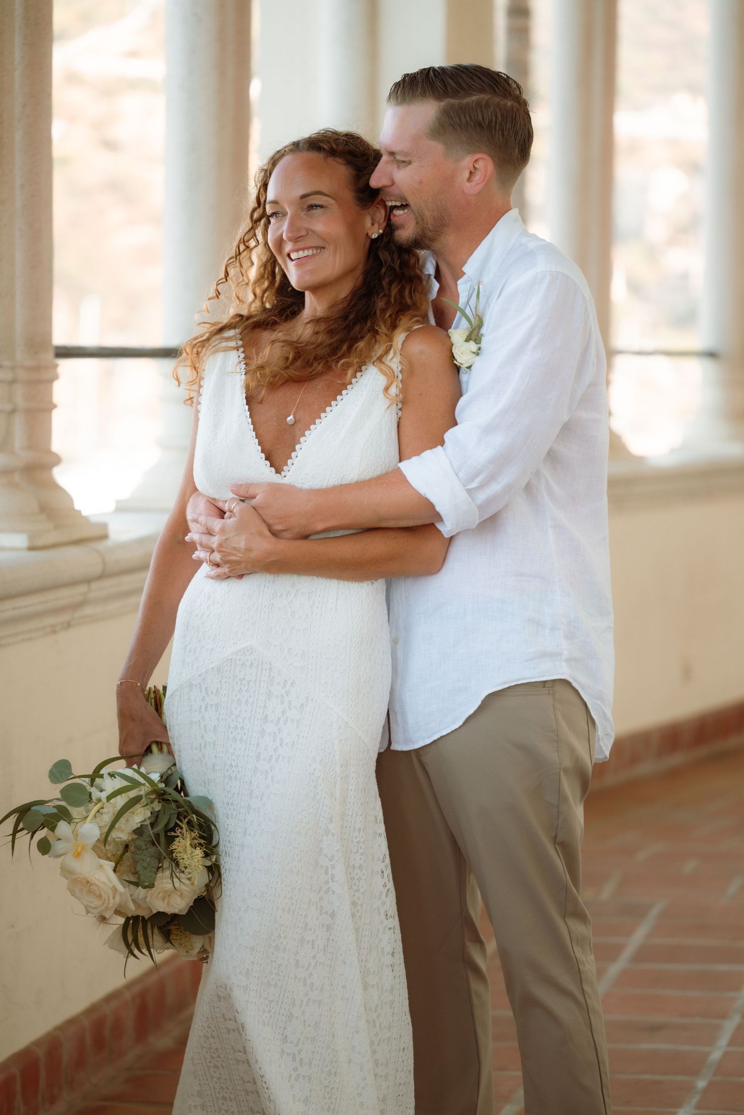 Bride in white lace gown holding bouquet embraced by groom in white shirt at elegant columned venue.