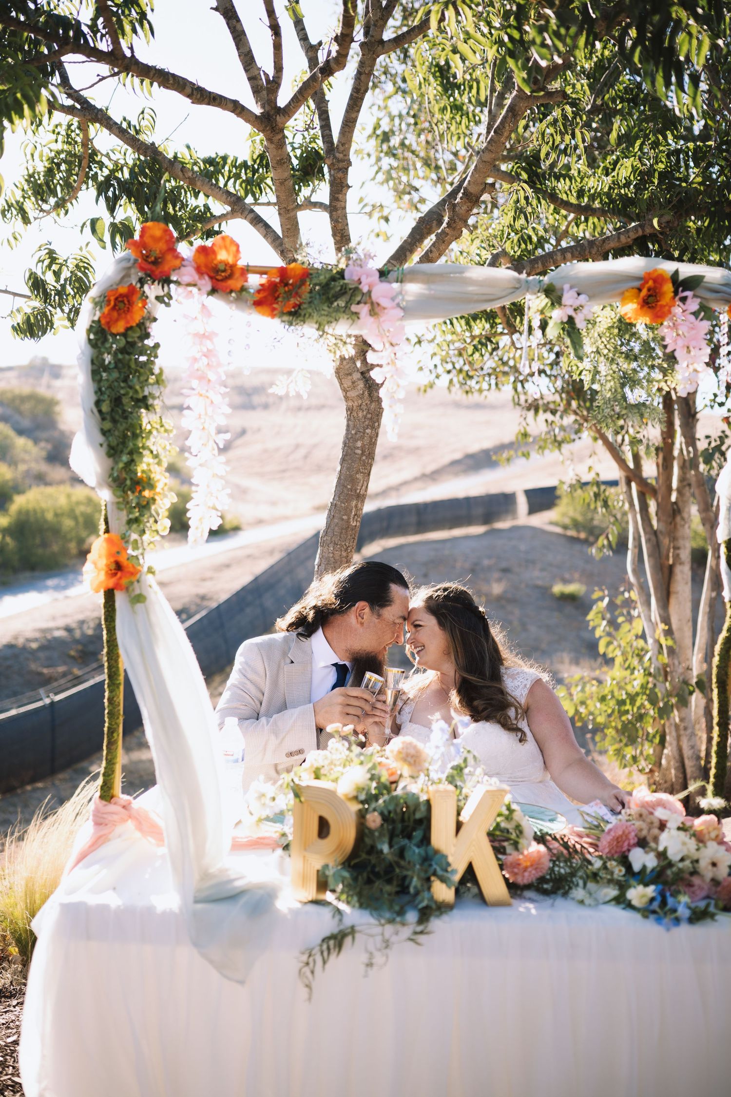 Bride and groom share a romantic moment at decorated sweetheart table under floral arch with P and K letters.