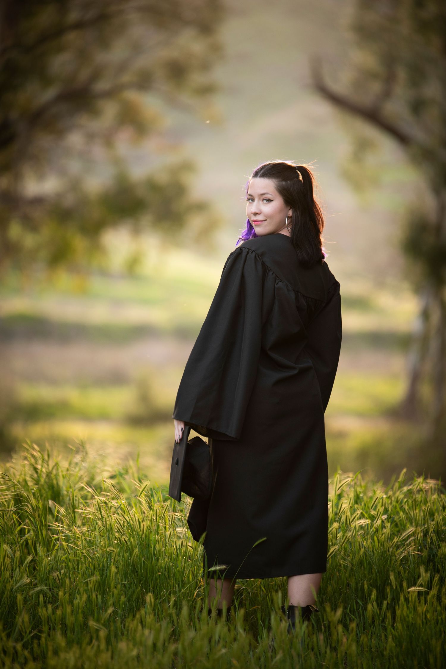 Woman in black graduation gown holding cap while standing in lush green field.