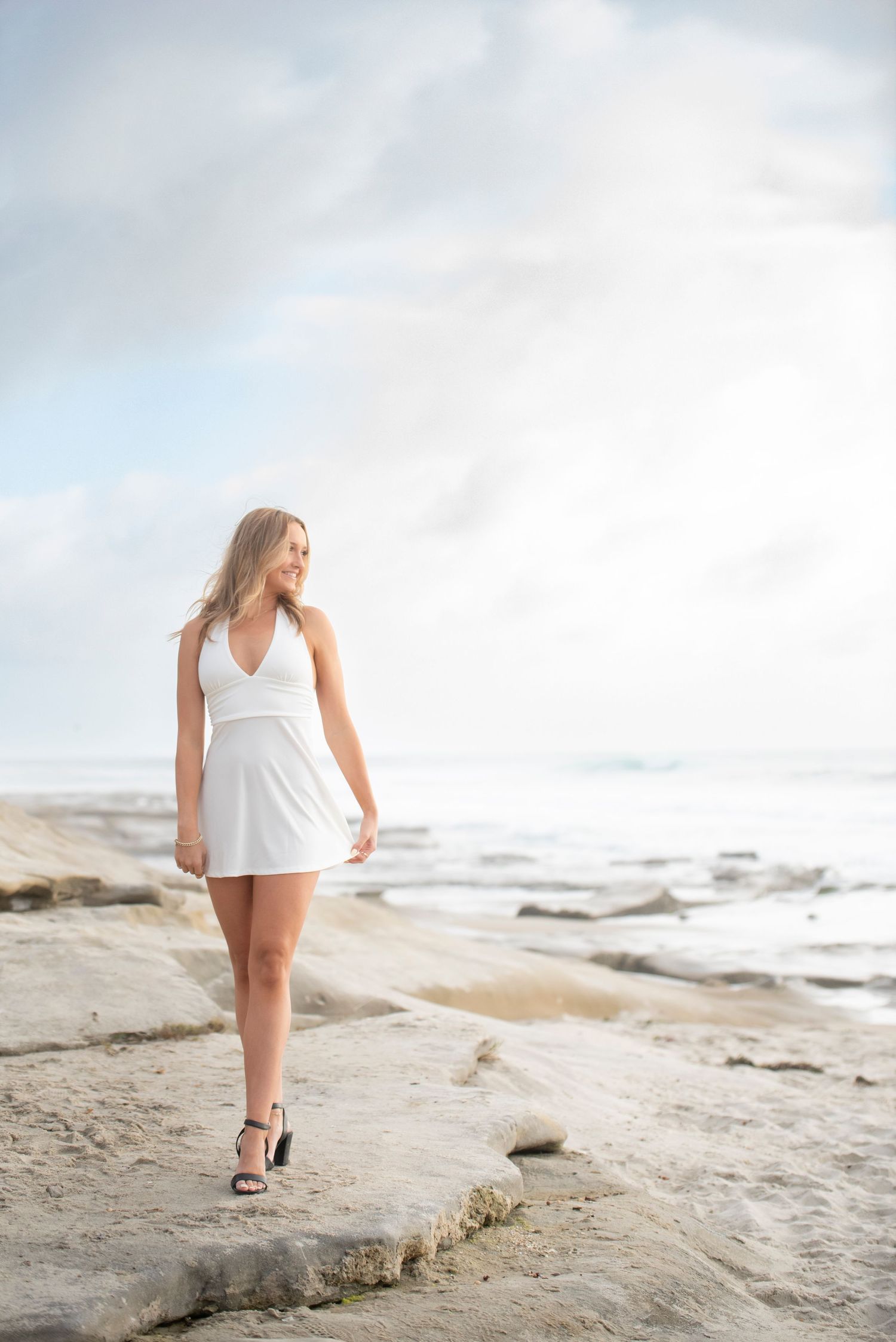 Young woman in white mini dress and heels standing on rocky beach shore with ocean and cloudy sky behind her.