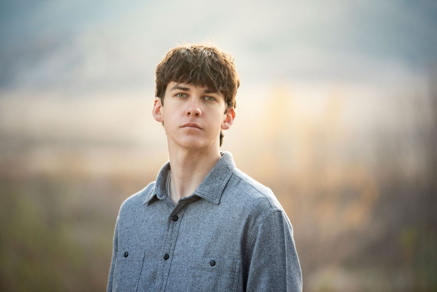 Young man in a gray button-up shirt standing outdoors with a blurred landscape background at dusk.