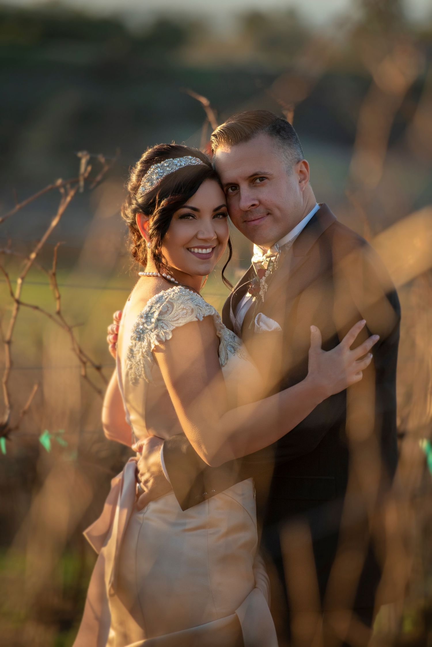 Smiling bride with jeweled headband embraces groom in warm golden sunset light among vineyard vines.