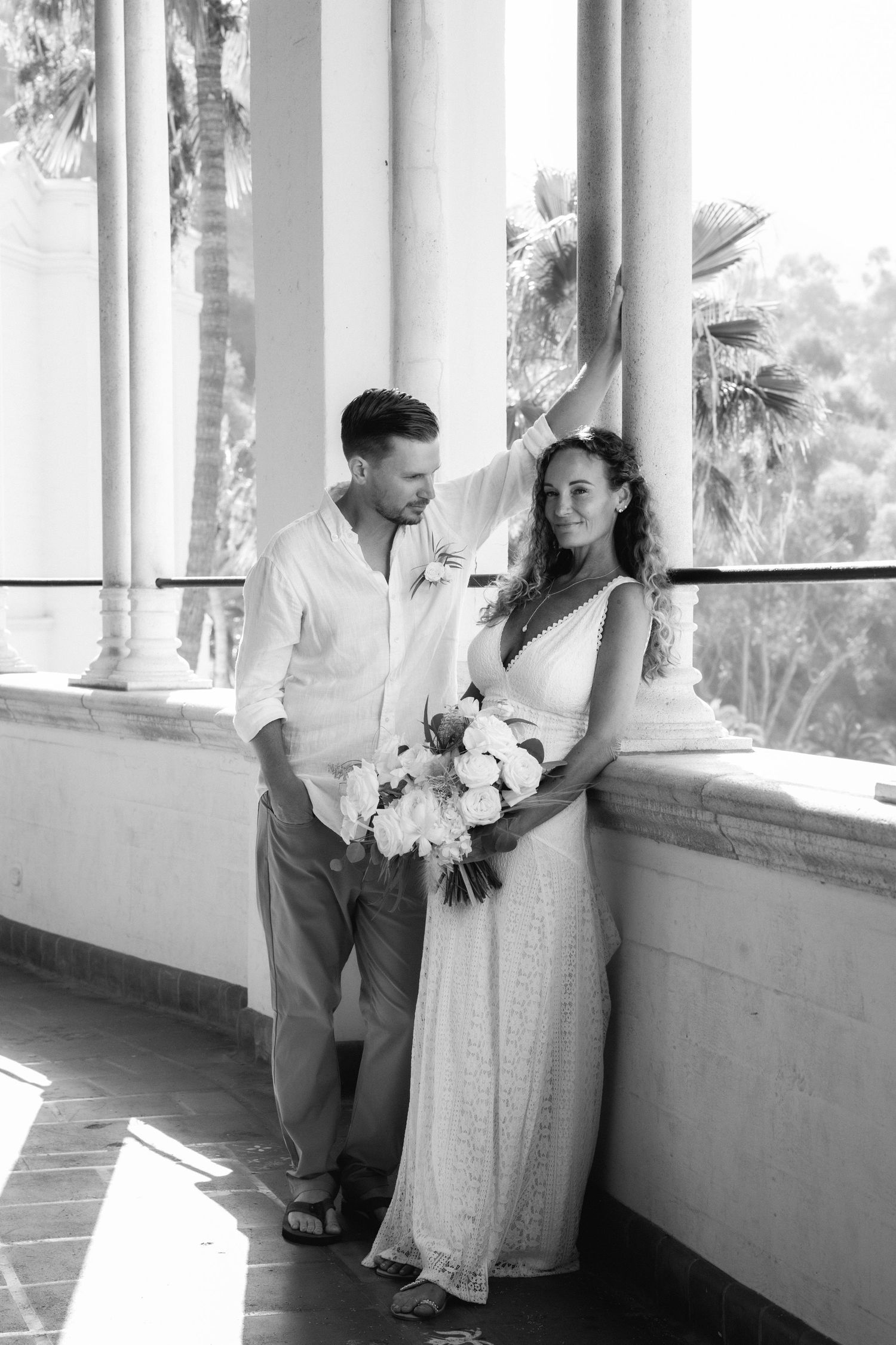 Black and white wedding photo of bride holding bouquet and groom standing together on a sunlit columned veranda with palm trees.