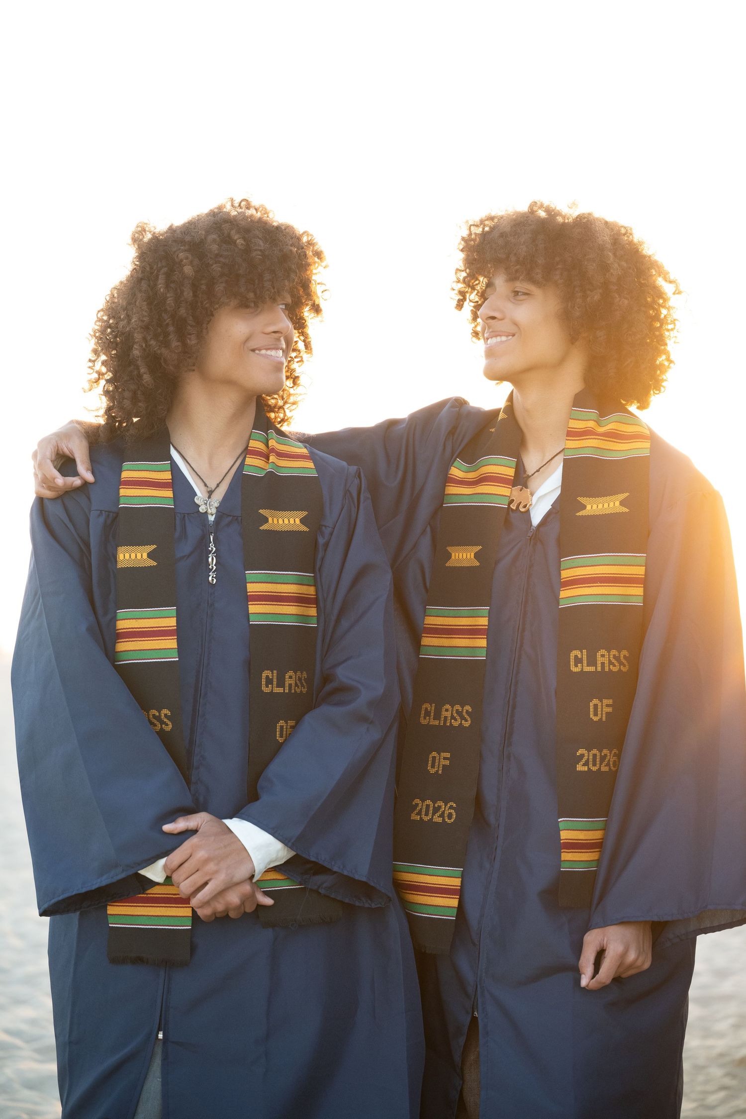 Twin graduates in matching blue gowns and kente stoles smile at each other on sunny beach.