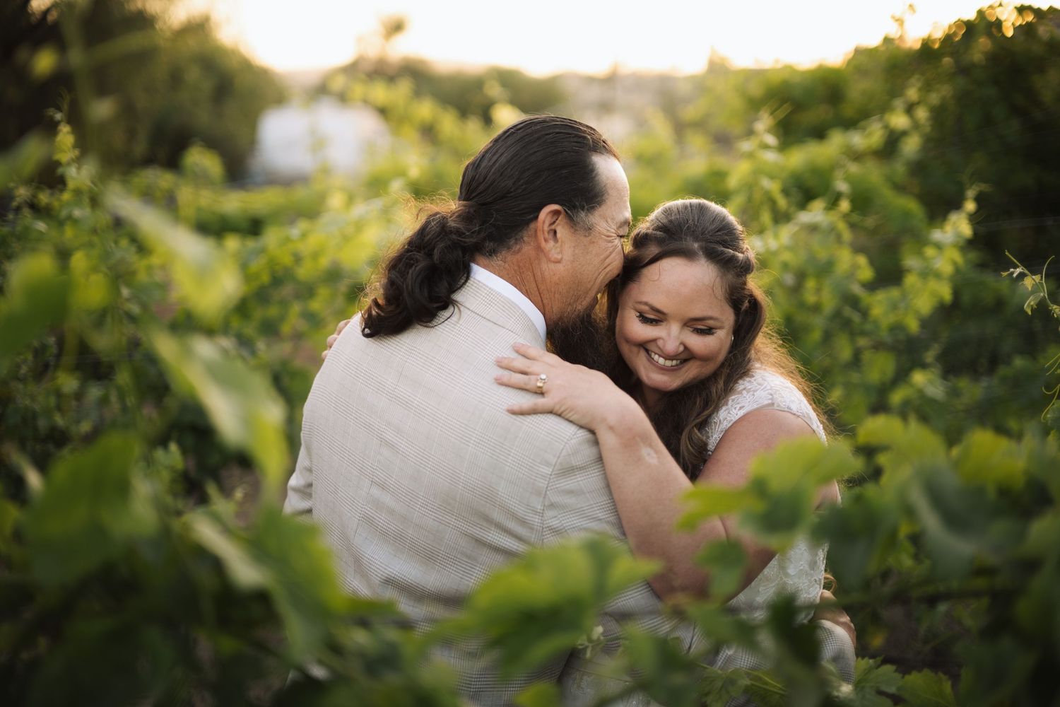 Bride smiles joyfully while embracing groom among lush green vineyard rows in warm golden sunset light.