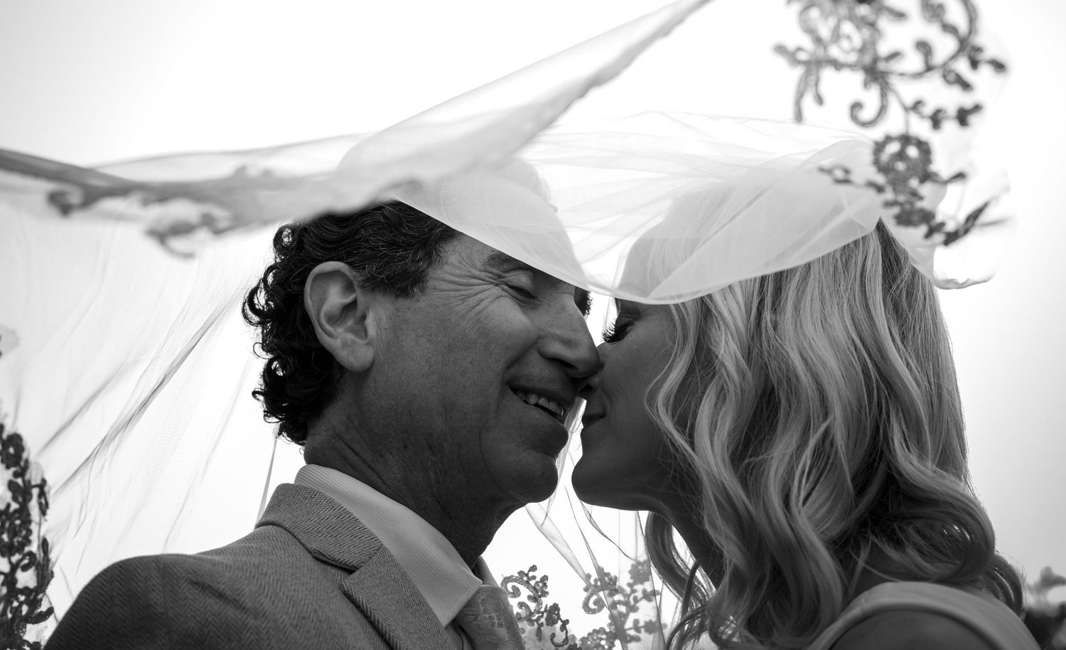 Bride and groom share a kiss under a veil in a classic black and white wedding photo.
