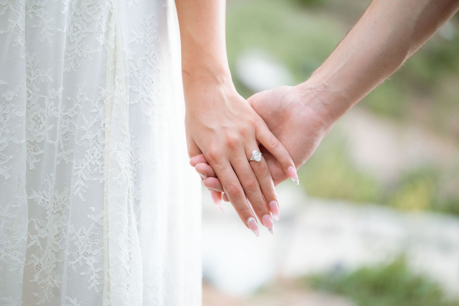Bride and groom holding hands, showcasing a diamond engagement ring, with the bride wearing a white lace wedding dress.