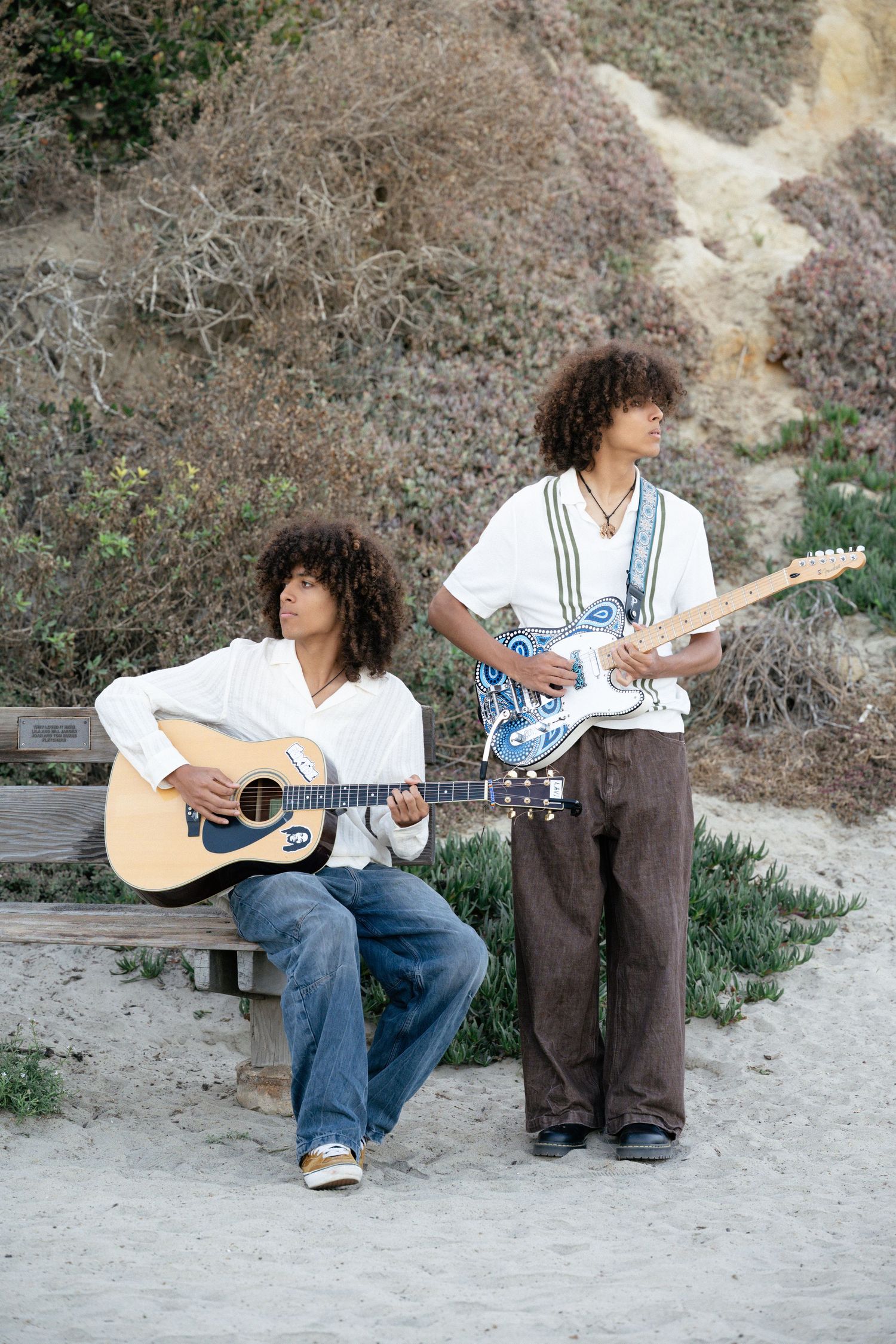 Twin brothers with curly hair play acoustic and electric guitars together on a sandy beach outdoor setting.