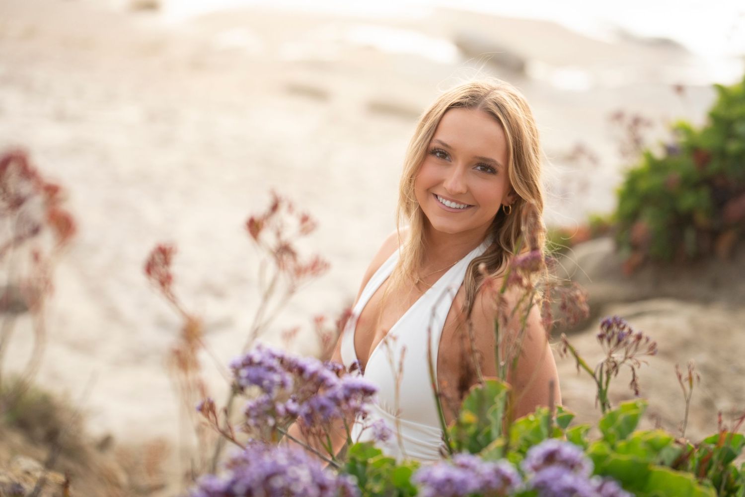 Smiling blonde woman in white dress surrounded by purple wildflowers and rocky coastal scenery.