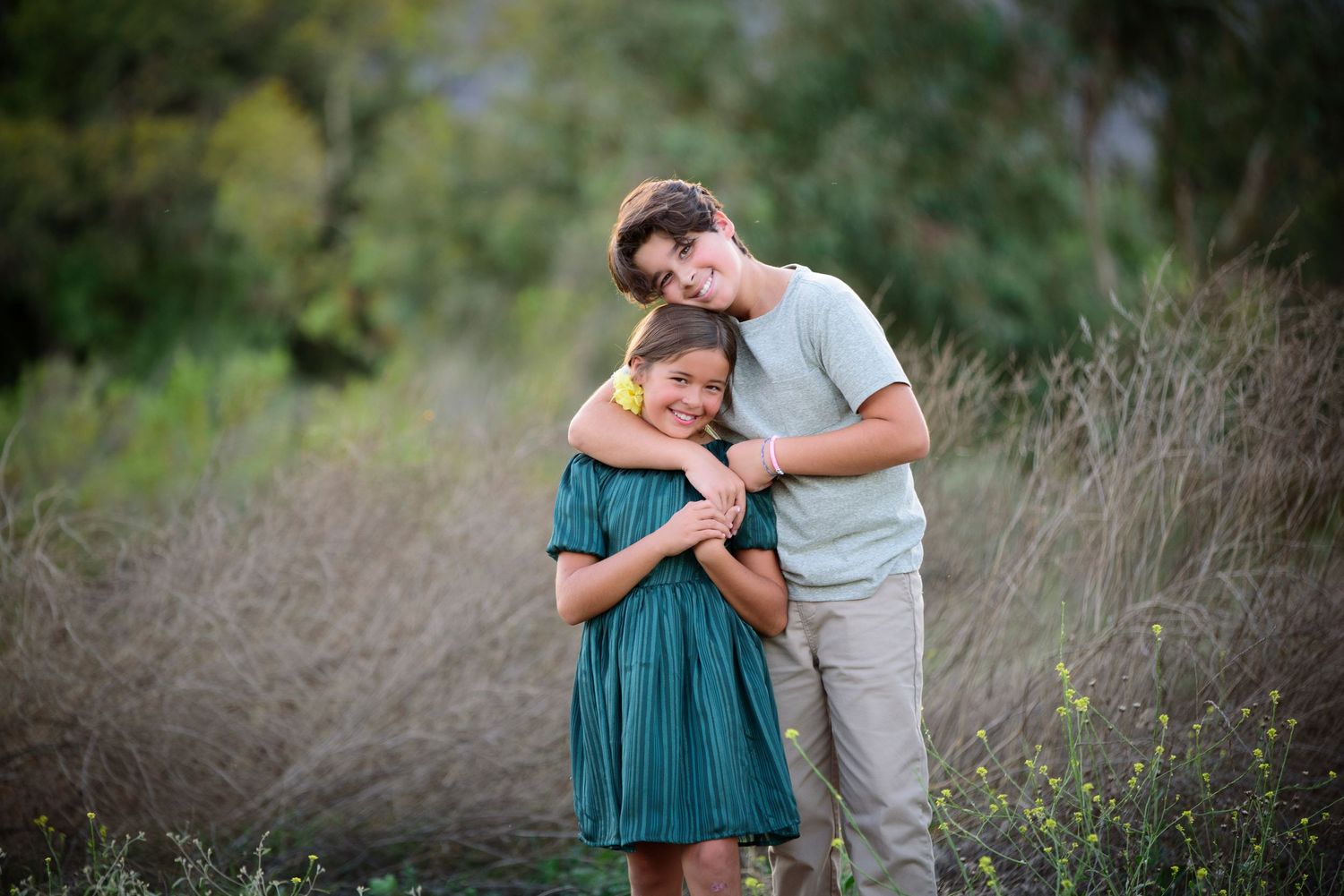 A teenage boy tenderly embraces a young girl in a teal dress along a nature trail at golden hour.
