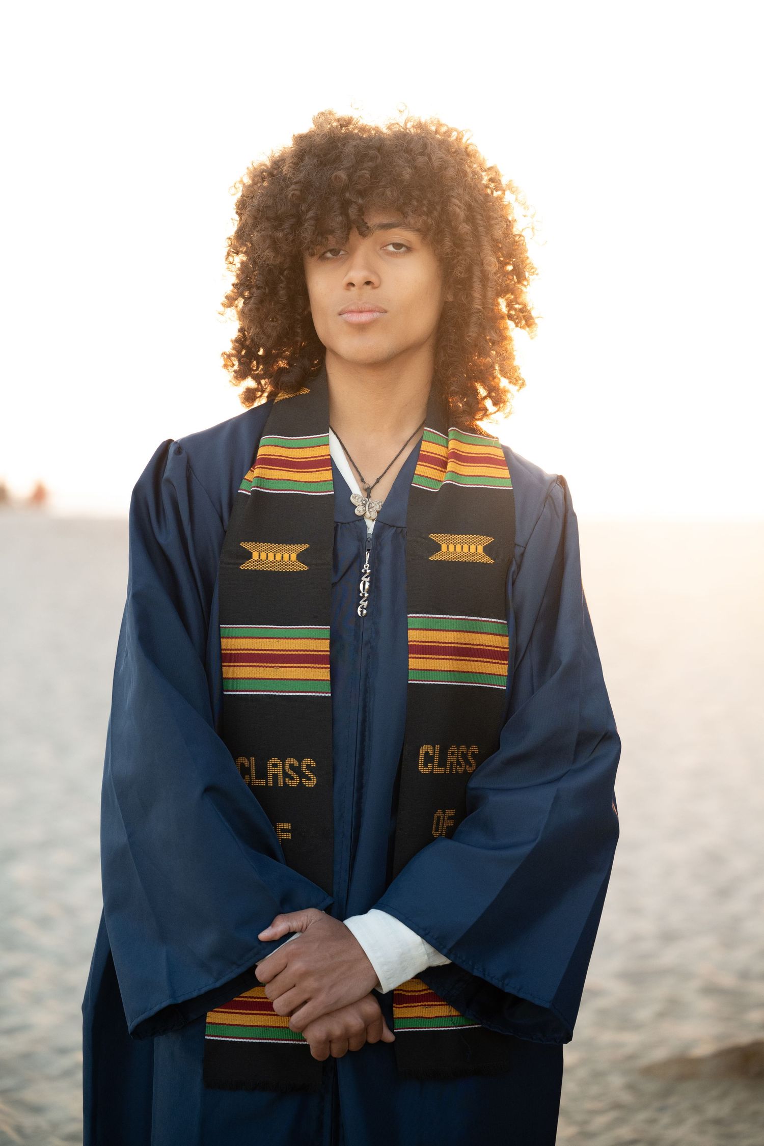 Young graduate wearing blue gown and kente cloth stole stands outdoors during golden hour.