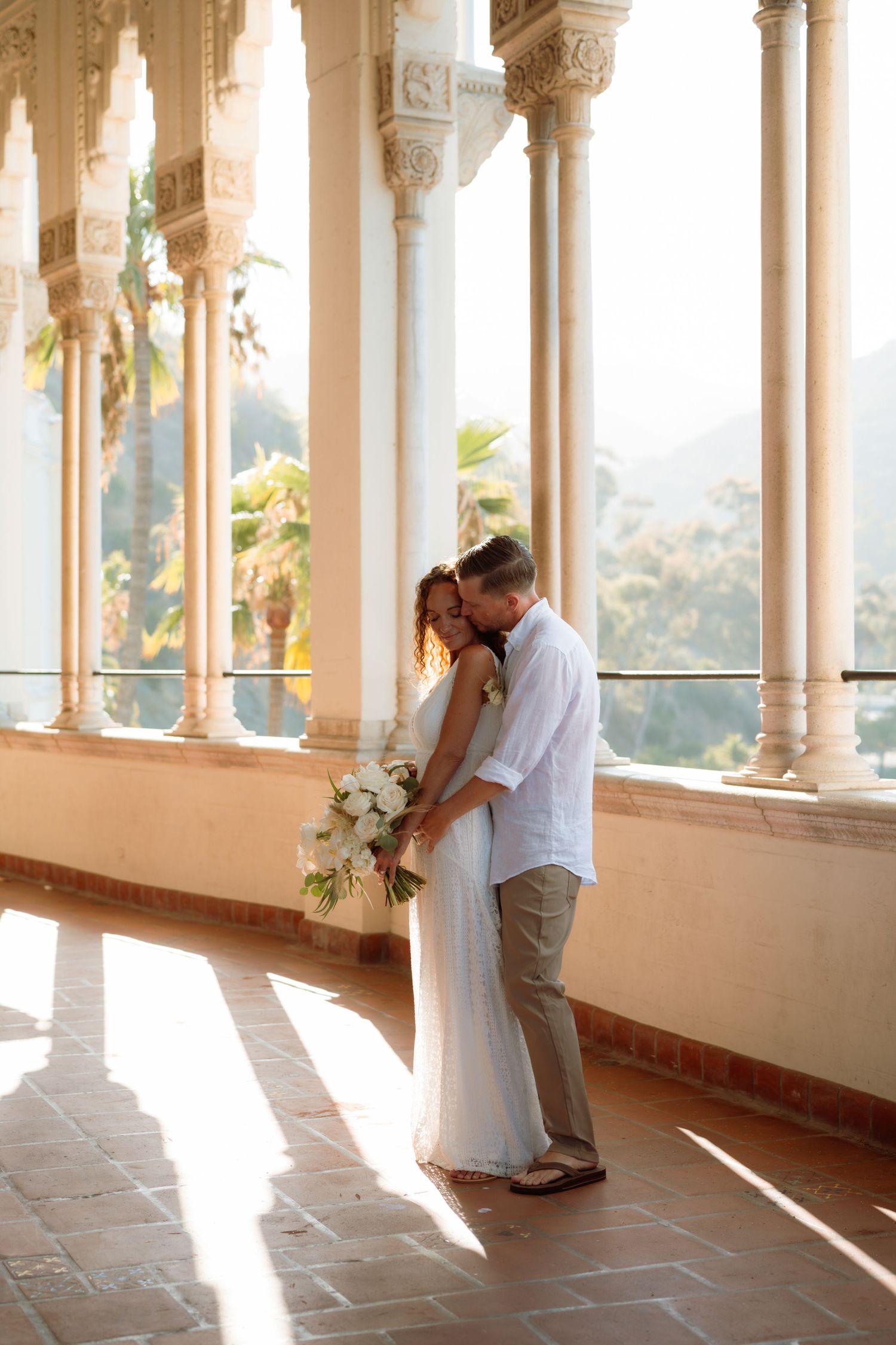 Couple sharing romantic kiss in sunlit colonial arcade with lush tropical garden backdrop on wedding day.
