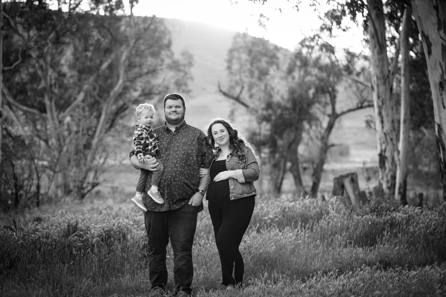 Black and white family photo of couple with toddler standing in a grassy field surrounded by eucalyptus trees at dusk.