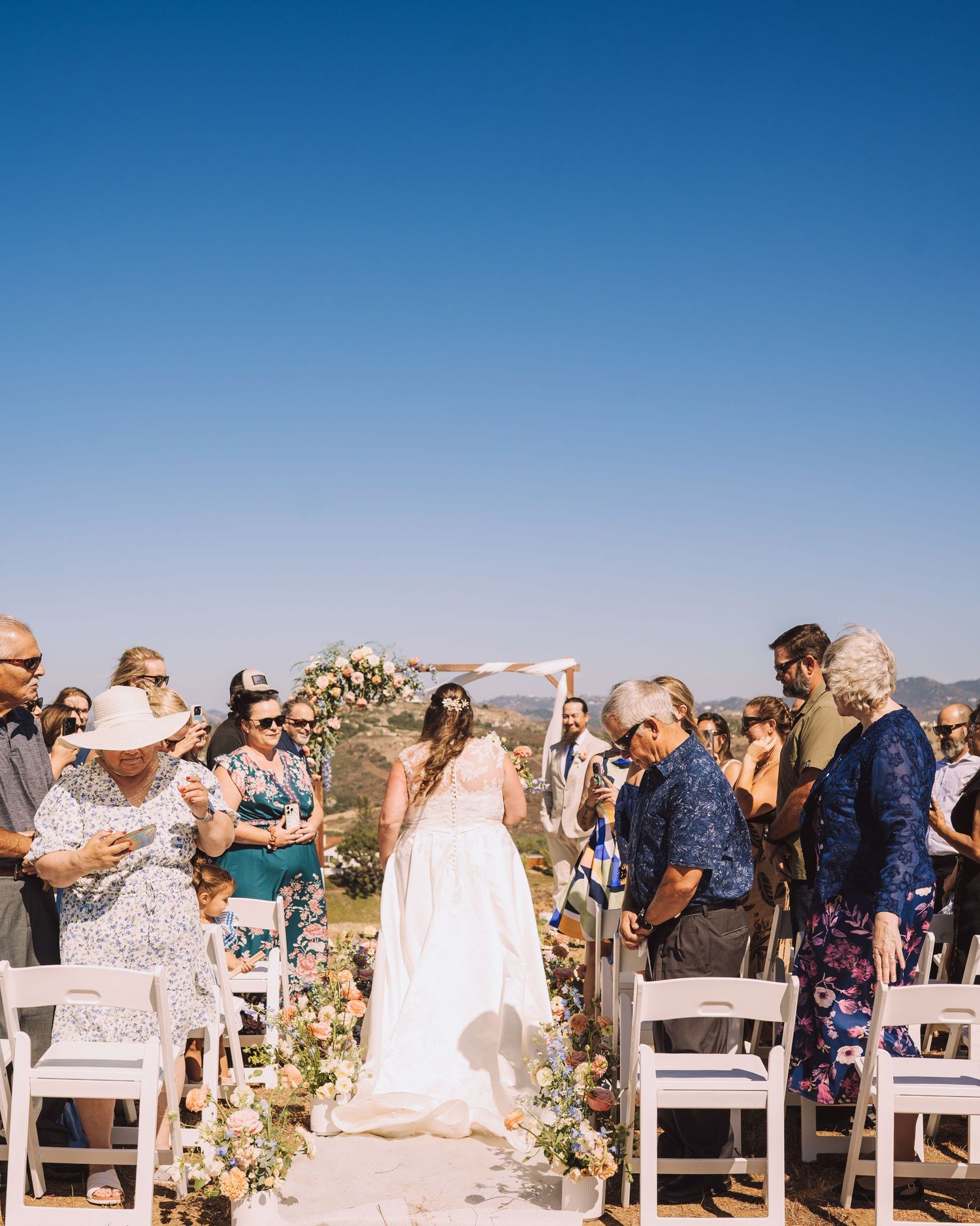 Bride walks down flower-lined aisle toward wooden arch at outdoor wedding ceremony with seated guests.