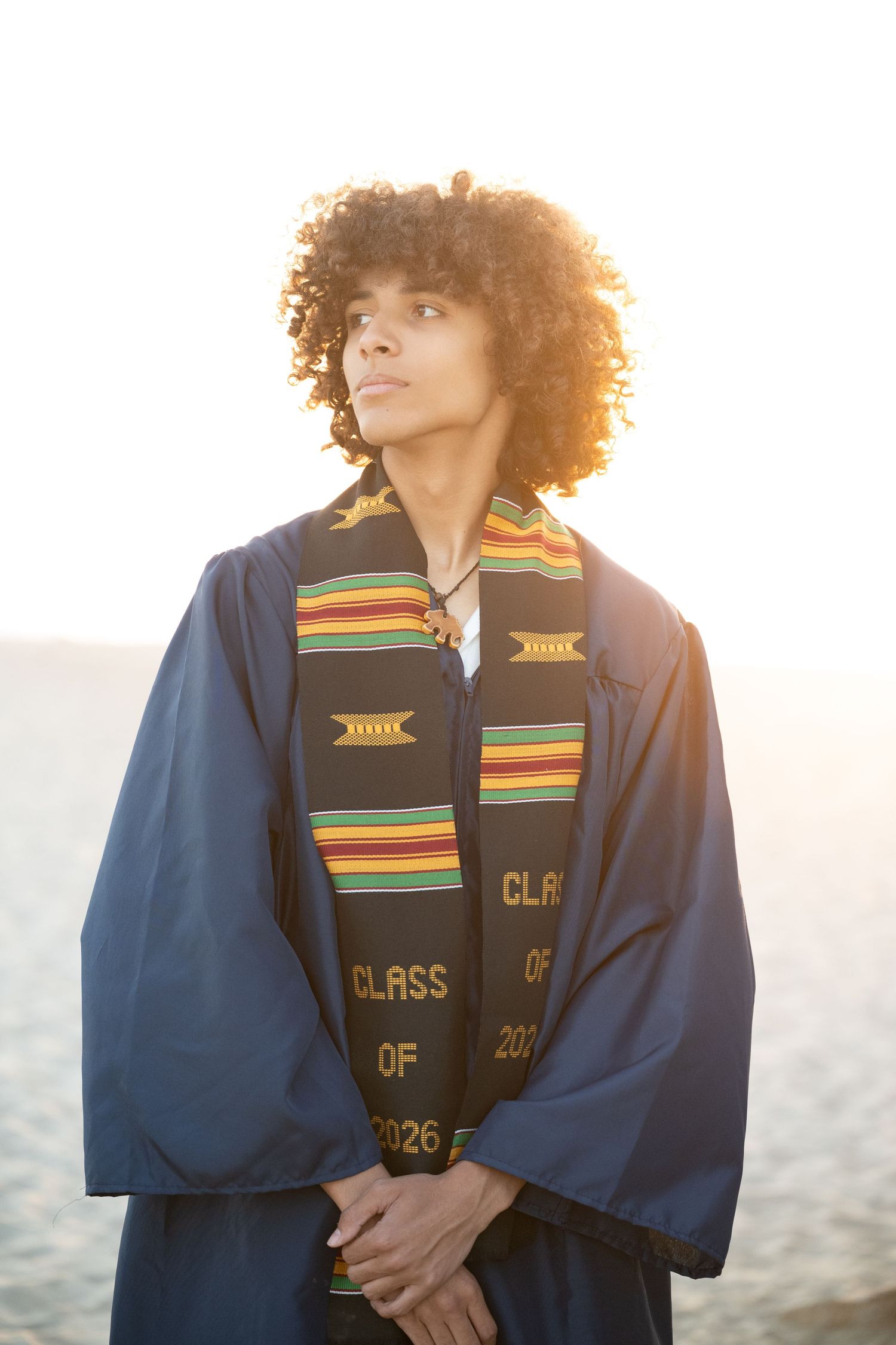 Graduate in blue cap and gown with colorful kente stole poses outdoors at sunset near ocean.