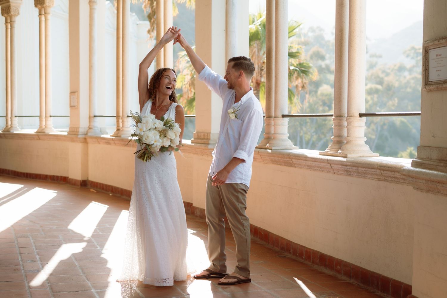 Bride and groom dancing on a sunlit balcony with white columns, bride holding a white bouquet in a flowing gown.