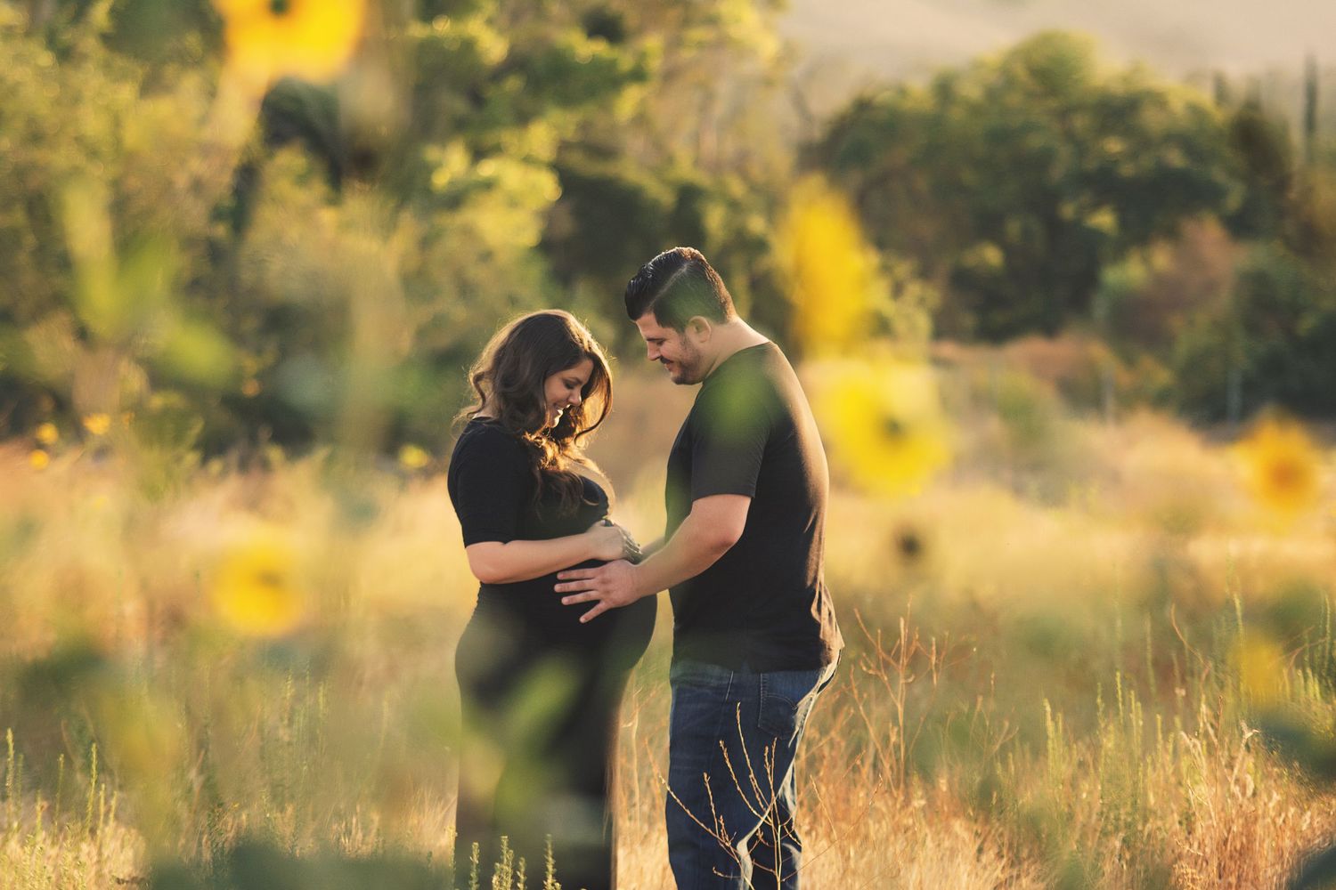 Couple sharing a tender maternity moment in a sunlit field with wildflowers and golden bokeh background.