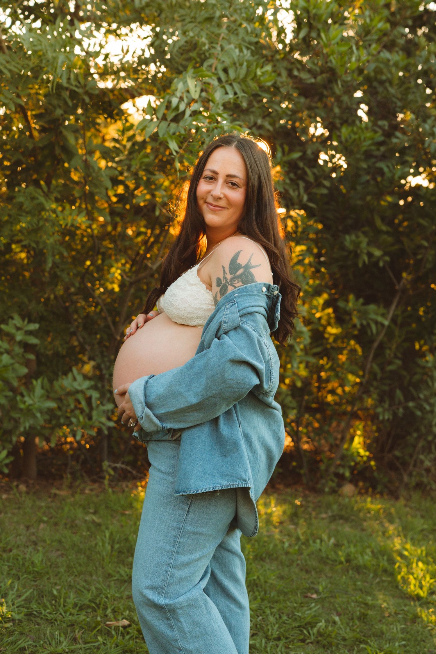Pregnant woman in denim jacket and jeans cradling baby bump outdoors at golden hour.