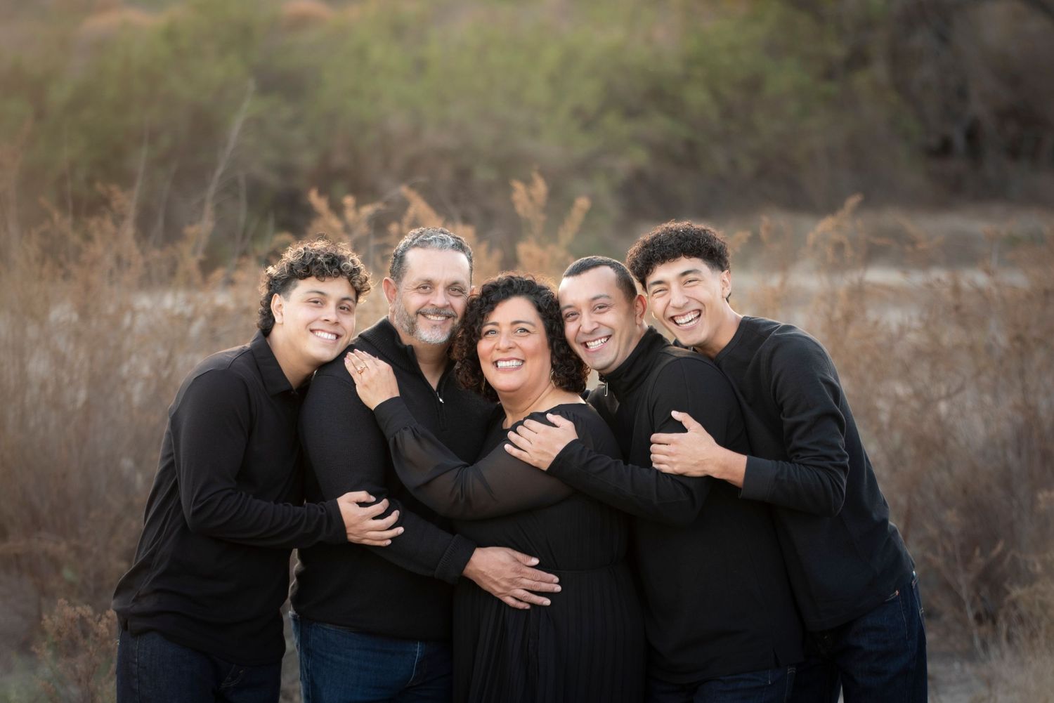 Family of five in black outfits embracing warmly in outdoor field.